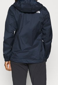 Vedenkestävä tummansininen takki, jossa on huppu, joustavat hihansuut ja tuuletusaukkoja. Takana on logo ja pinta on sileä ja kiiltävä.