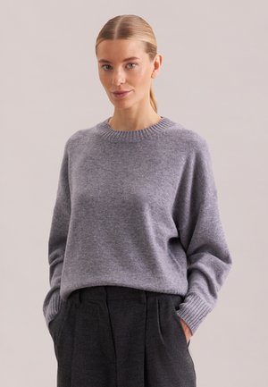 Frau mit zurückgebundenen blonden Haaren, die einen hellgrauen Pullover und dunkelgraue Hosen trägt, mit den Händen in den Taschen, neutraler Hintergrund.