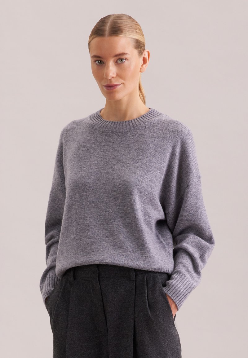 Femme aux cheveux blonds attachés en arrière, portant un pull gris clair et un pantalon gris foncé, les mains dans les poches, fond neutre.