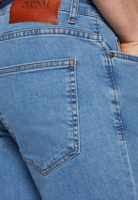 Világoskék farmernadrág narancssárga varrással, melyen egy "2YDNM" feliratú bőr címke található, és egy geometrikus mintázatú hátsó zseb.