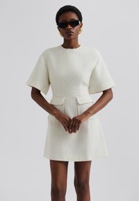 Robe à manches courtes de couleur crème, faite d'un tissu texturé, avec deux poches avant et une taille cintrée, dotée d'une jupe droite.
