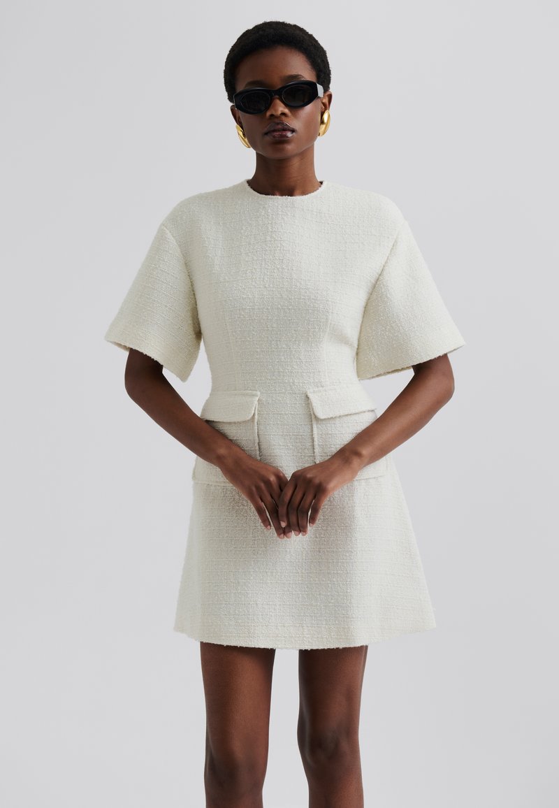 Robe à manches courtes de couleur crème, faite d'un tissu texturé, avec deux poches avant et une taille cintrée, dotée d'une jupe droite.