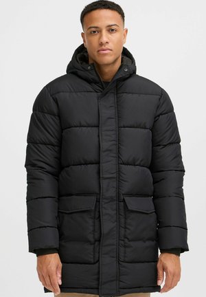 Manteau rembourré noir avec capuche, présentant un design matelassé, deux poches avant et une fermeture zippée. Texture lisse, longueur au genou.