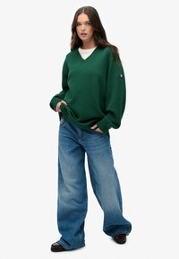 Groene oversized gebreide trui met een V-hals, geribde mouwen en zoom. Gecombineerd met loszittende blauwe spijkerbroeken en zwarte schoenen.