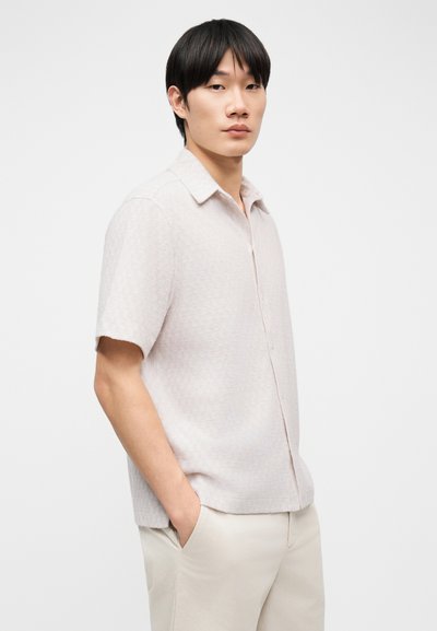 Νέος άνδρας με μαύρα μαλλιά στέκεται με τα χέρια στις τσέπες, φορώντας ανοιχτό μπεζ κοντομάνικο υφασμένο πουκάμισο και παντελόνι που ταιριάζουν, σε ουδέτερο φόντο.