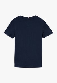 Camiseta de manga corta en azul marino con cuello redondo, con un pequeño lazo de tela rojo, blanco y azul en la parte posterior del cuello, diseño liso.