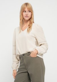 Beige V-kaula-aukollinen pusero, jossa on pitkät, pussimaiset hihat, joissa on kirjailtu koristeita kaltereissa; yhdistetty oliivinvihreisiin housuihin.