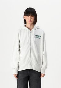 Vaaleanharmaa vetoketjullinen huppari, jossa on nyörit ja vihreä logo rinnassa. Pehmeää materiaalia, löysä istuvuus ja kengurupussit.