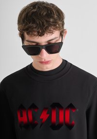 Zwarte sweatshirt met een gedurfde AC/DC-logo in een verloop van rood. Model draagt hoekige zwarte zonnebril en heeft een zachte, afgeronde halslijn.