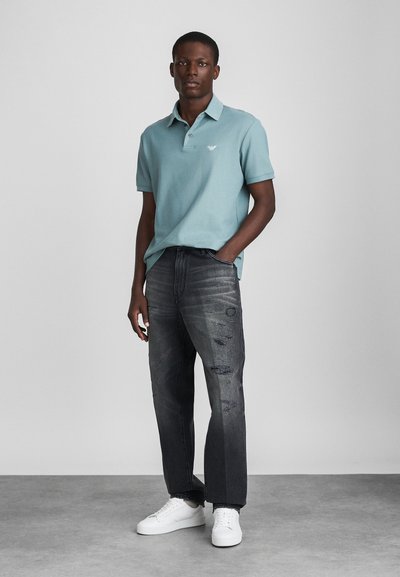 Svetlomodrá polokošeľa s krátkymi rukávmi, šedé poškodené džínsy a biele tenisky. Jemné logo na polokošeli a viditeľná textúra látky.