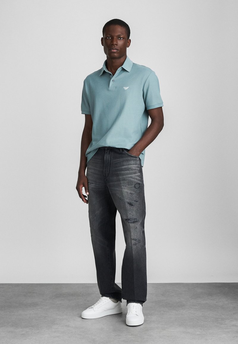 Polo bleu clair à manches courtes, jeans gris déchirés et baskets blanches. Logo discret sur le polo et texture du tissu visible.