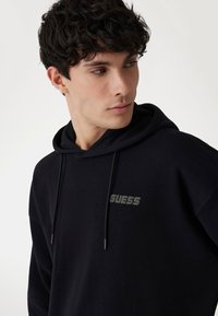 Fekete pamut kapucnis felső, húzózsinórral, a mellkasán egy szürke "GUESS" logóval. Sima textúrájú és laza szabású dizájn.