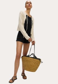 Giacca corta in tessuto cremoso, tuta nera, sandali neri con cinturini e una grande borsa rotonda in paglia intrecciata con manici neri.