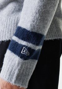 Grijze trui met een geribbelde textuur en marineblauwe en lichtgrijze strepen op de mouwen. Inclusief een klein geborduurd logo op de manchette.