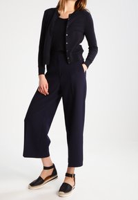 Kvinna som bär en svart knappad cardigan, svart topp, breda cropped marinblå byxor och svarta espadriller med ankelsnodd som står.