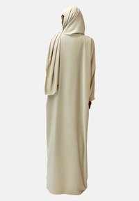 Lång crèmefärgad abaya med ett flödande mönster, som har en lös passform, långa ärmar och en matchande hijab. Slät tygtextur.