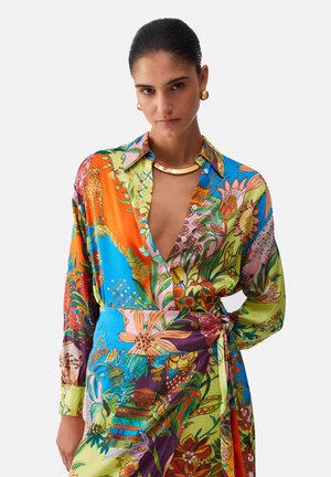 Femme aux cheveux plaqués en arrière porte une robe portefeuille éclatante à motifs floraux et animaliers avec un décolleté en V profond, ainsi que des boucles d'oreilles créoles dorées et un collier.