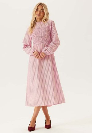 Vrouw met lang blond haar, gekleed in een roze, jurk met lange mouwen en enkel lengte met verticale strepen en bordeauxrode hakken, staand met samengevouwen handen.