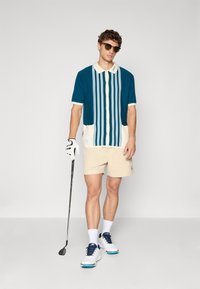 Mann som har på seg solbriller, stripete kortermet skjorte, beige shorts, hvit golfhanske og sko, som holder en golfklubbe mot en ensfarget bakgrunn.