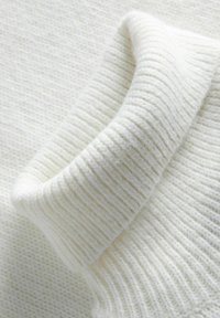 Pull en tricot blanc avec un col à côtes et un tissu doux et texturé. Le motif de tricot est serré, créant une apparence chaleureuse.