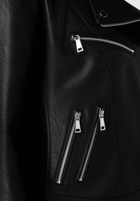 Gros plan sur une veste en cuir noir montrant trois poches zippées verticales argentées et un bouton pression sur le rabat du col.
