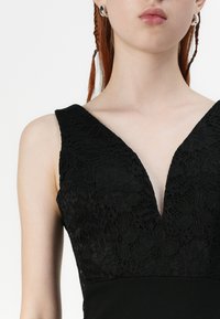 Musta mekko, jossa on kukkainen pitsikuvio bodice syvällä V-aukolla, sileä kangas alapuolella ja leveät olkaimet. Hopeiset renkaat ovat näkyvissä.