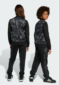 Černá a šedá maskáčová sada bundy a kalhot s žebrovanými manžetami a pasem, s černými bočními pruhy a texturovanými materiály.