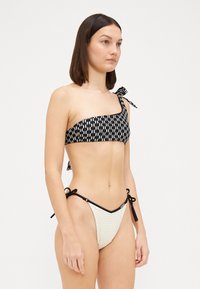 Bikini zgornji del v črno-beli vzorčasti zasnovi z enim naramnicom in stranskimi zavezavami, v kombinaciji s smetanovimi nizkimi hlačkami, ki imajo črne obrobe in resice.
