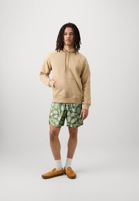 Sweatshirt beige à capuche avec une poche avant, associé à un short vert à motifs avec des motifs de feuilles, et des chaussures slip-on marron avec des chaussettes blanches.