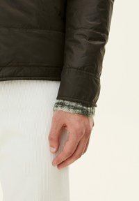 Dark green puffer jacket with stitch detailing on the sleeve, over a white ribbed texture, featuring a knitted cuff in green and gray.