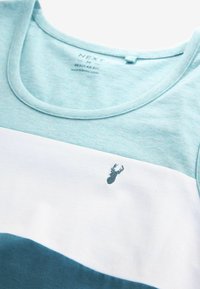 Vaaleansininen, valkoinen ja tumman teal-vihreä väriblokattu paita, jossa on pieni brodeerattu tumman teal-vihreä toimittajan sarven logo rinnassa.