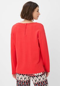 Felpa rossa con maniche lunghe, scollatura rotonda e dettaglio con zip sulla schiena. Tessuto morbido e spacco laterale. Abbinata a pantaloni con fantasia.