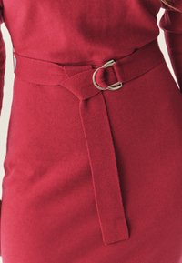 Rød strikket kjole med detaljer, der inkluderer et matchende stofbælte med en sølvmetalring, der fremviser en glat tekstur og tætsiddende design.