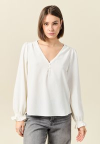Blusa bianca con scollatura a V e tre piccoli bottoni sul petto. Presenta maniche lunghe con polsini arricciati e un tessuto liscio e leggero.
