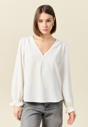 Witte blouse met een V-hals en drie kleine knopen op de borst. Heeft lange mouwen met geplooide manchetten en een gladde, lichte stof.