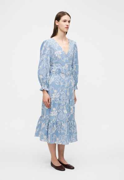 Femme debout, portant une robe midi bleu clair à manches longues avec une broderie blanche sur le thème de la mer et des chaussures plates foncées sur un fond blanc.
