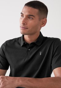 Polo negro de mezcla de algodón con collar, tapeta de dos botones y logo bordado en el pecho. Textura suave, diseño ajustado.