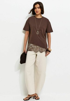 Mujer con una blusa marrón de manga corta con ribete de encaje, pantalones anchos color crema, sandalias negras con tacón, collar dorado, pulseras y sosteniendo un bolso tejido oscuro.