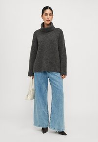 Maglione lavorato a maglia grigio scuro con collo alto, abbinato a jeans a gamba larga di un blu chiaro. Il modello tiene in mano una borsa bianca, in piedi su uno sfondo neutro.
