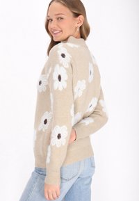 Pulover bej cu model de flori albe și centre întunecate. Guler și manșete cu tiv rișat. Textură moale cu design ajustat. Purtați-l cu blugi albastre.