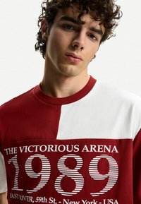 T-shirt in cotone rosso e bianco con testo in grassetto "L'ARENA VITTORIOSA 1989" e righe orizzontali. Collo rotondo e maniche corte.