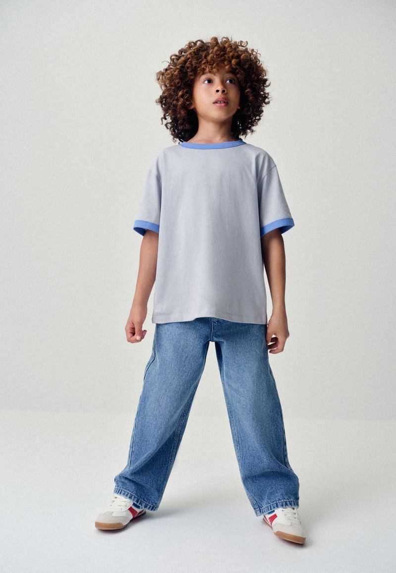 Child with curly hair wearing a light gray t-shirt with blue trim, loose blue jeans, and white sneakers standing with legs apart on a plain background.