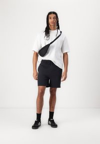 Black sling bag, oversized white shirt, dark shorts, black sneakers with white stripes, standing on a neutral background.