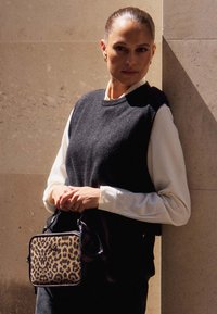 Kabelka přes rameno s leopardím potiskem a černými akcenty, obdélníkového tvaru, texturovaná látka, se zapínáním na zip, kterou drží osoba v černém svetru.