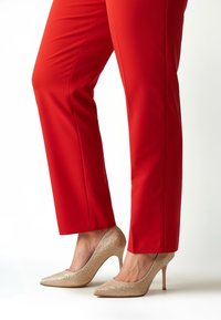 Rote eng anliegende Hose kombiniert mit schimmernden goldfarbenen Pumps mit hohem Absatz, die eine spitze Zehenpartie und eine strukturierte, glitzernde Oberfläche aufweisen.