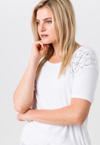 Korte mouw witte T-shirt met decoratieve zilveren accenten op de schouders, ronde halslijn en een zachte stoftextuur.