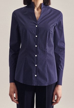 Kvinde iført navyblå skjorte med knapper og et lille hvidt ternet mønster samt lange ærmer, kombineret med mørke bukser.