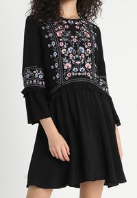Vestido negro con bordados florales en rosa y azul. Presenta mangas largas acampanadas y un dobladillo con volantes. Hecho de tela ligera.