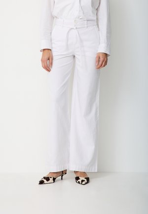 Frau trägt weiße, weit geschnittene Hosen mit gebundenem Gürtel, weißes, hineingestecktes Hemd und schwarz-weiß gemusterte Pumps mit kleinem Absatz.