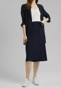 Marinblå blazer över en vit topp med spetsdetaljer, kombinerad med en knälång marinblå kjol som är knutad i midjan, och vita sneakers. Slät tygtextur.